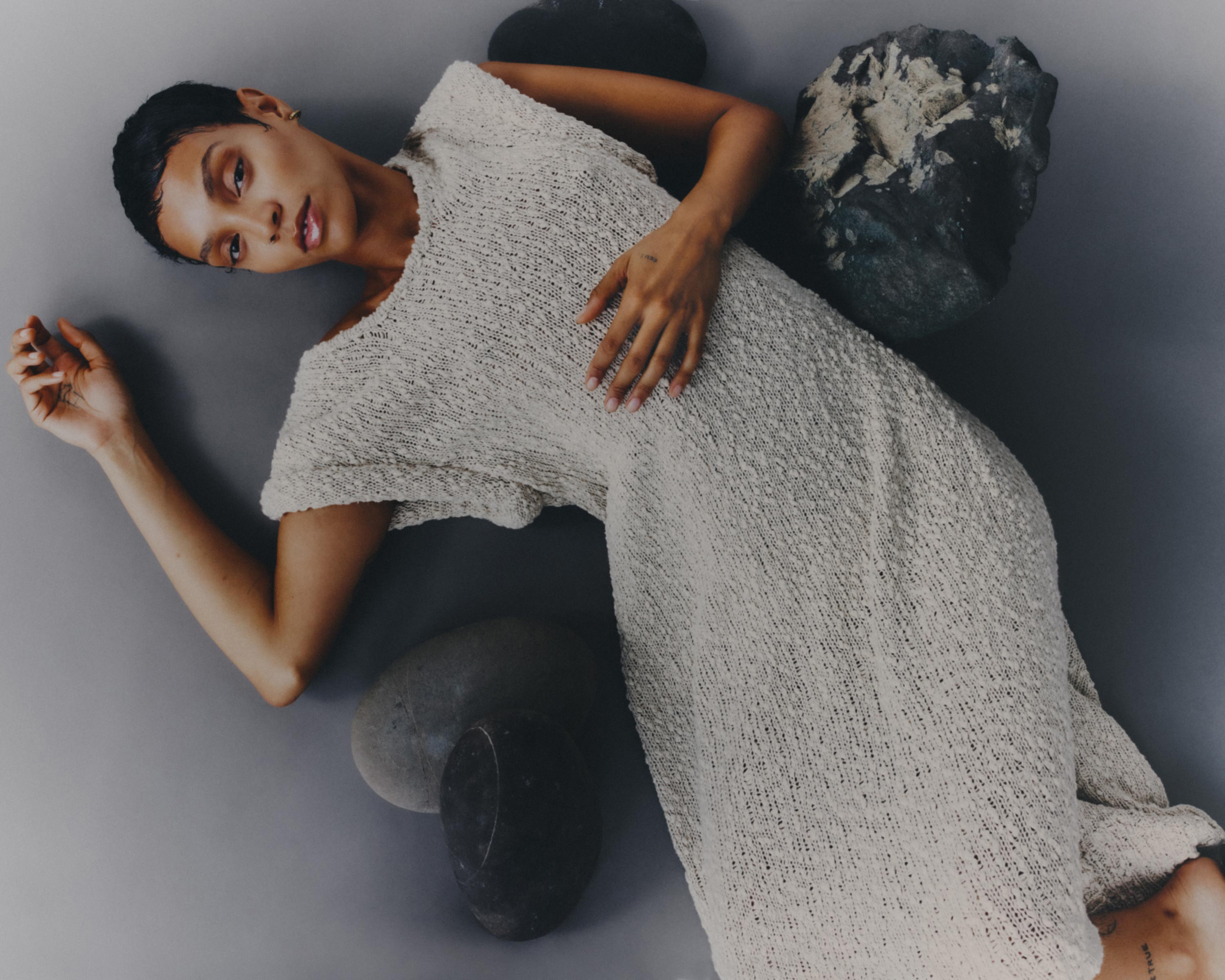 Adult woman in light dress lies on a gray surface, posed with rocks. Studio shot with neutral color palette, fashion or beauty editorial style.