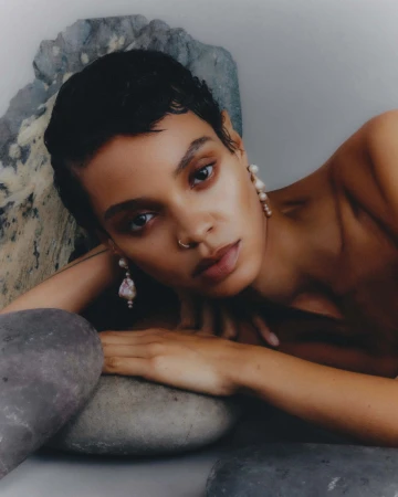 Portrait of a woman with short dark hair reclining near grey rocks wearing earrings, shot with natural light and shallow depth of field.