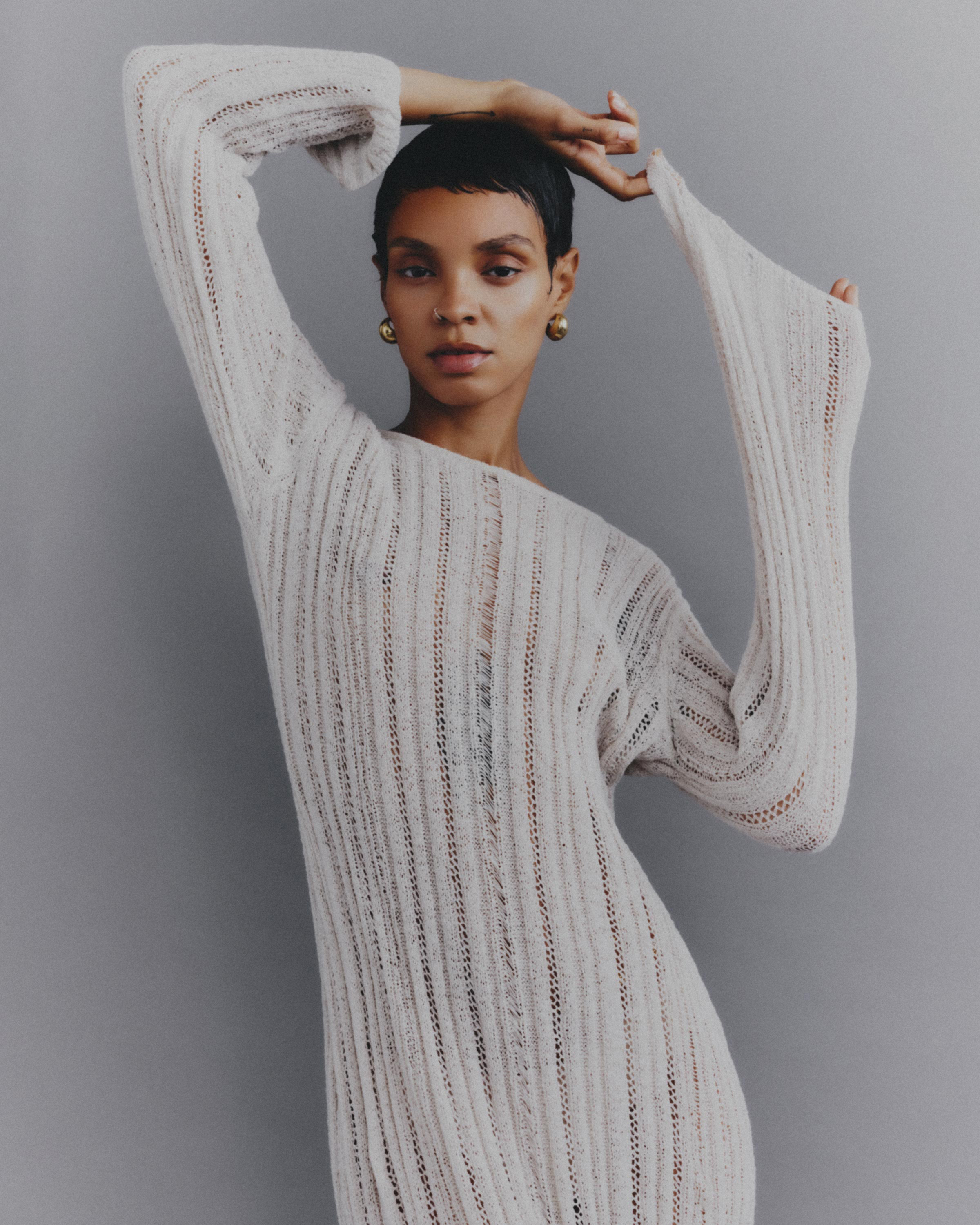 Portrait of an adult woman with short dark hair wearing an off-white knit dress against a grey background, with her arms raised.