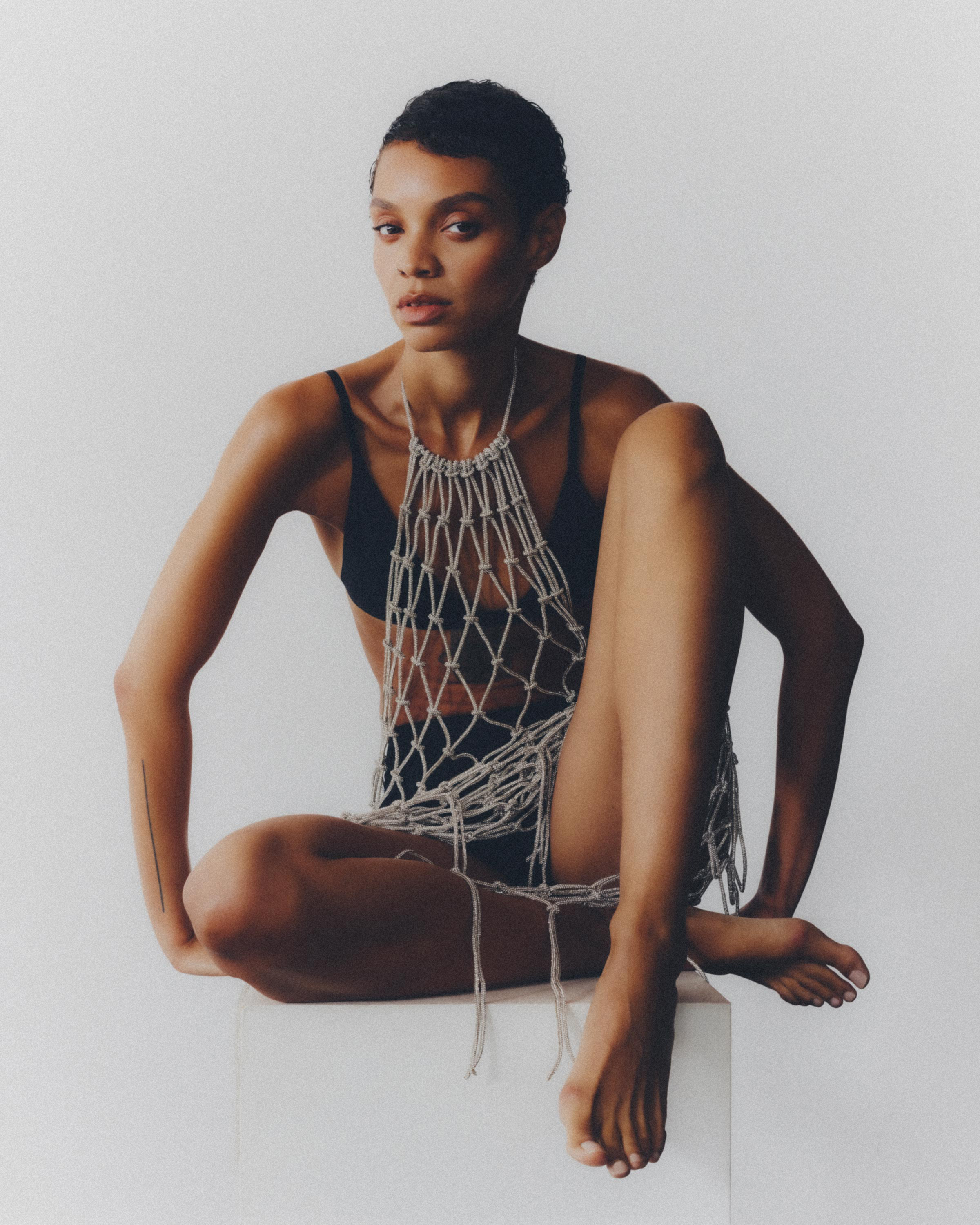 Adult woman with short hair poses indoors wearing a black bra and a crocheted net garment, sitting on a white box with soft lighting.