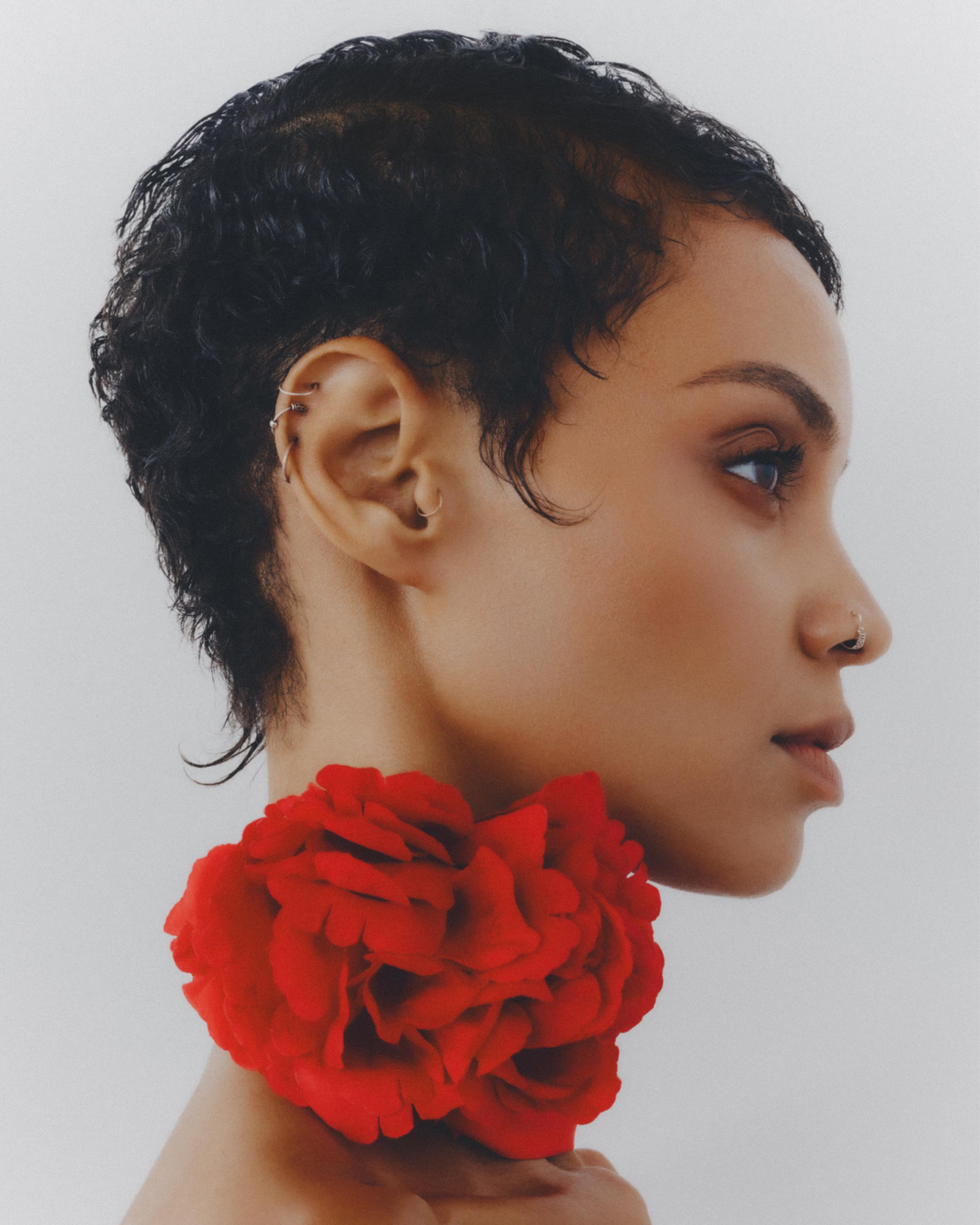 Profile portrait of a woman with short curly hair and nose ring, adorned with a large red rose, set against a soft white backdrop.