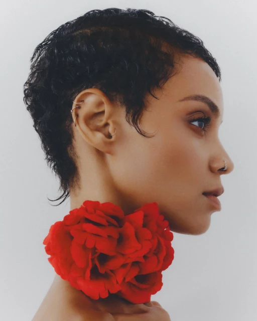Profile portrait of a woman with short curly hair and nose ring, adorned with a large red rose, set against a soft white backdrop.