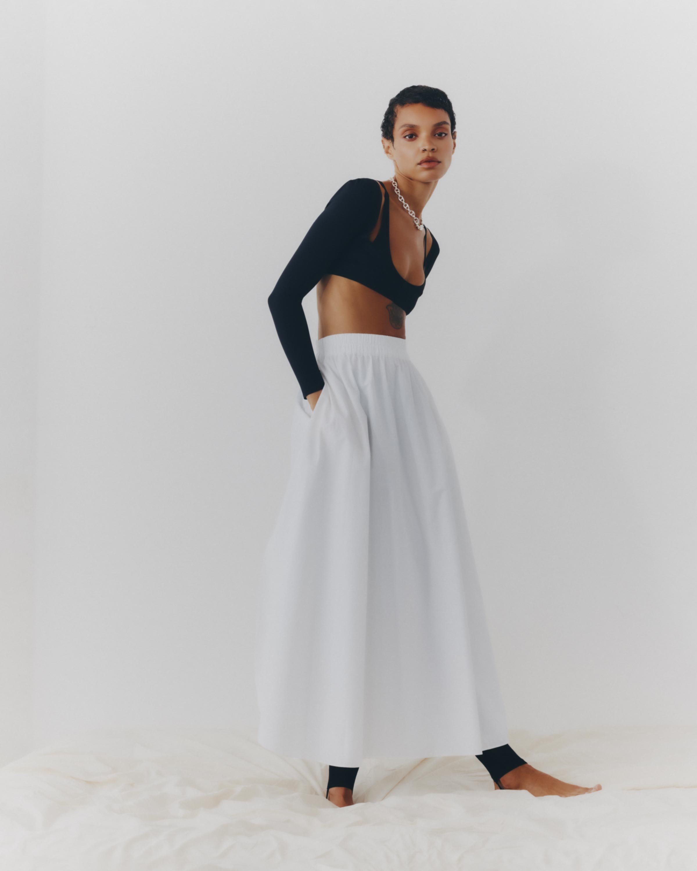 Full-length portrait of an adult female model wearing a black long-sleeve crop top and a large white A-line skirt in a photo studio.