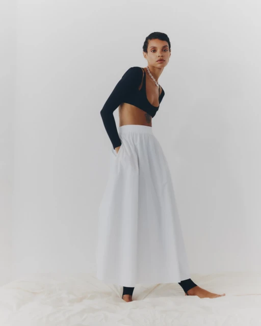 Full-length portrait of an adult female model wearing a black long-sleeve crop top and a large white A-line skirt in a photo studio.