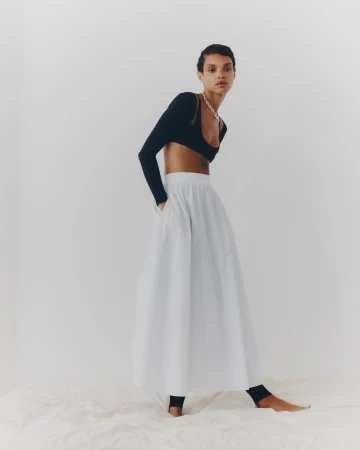 Full-length portrait of an adult female model wearing a black long-sleeve crop top and a large white A-line skirt in a photo studio.