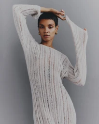 Model Akira Reid posing with raised arms wearing a textured off-white knit dress against a solid grey backdrop.