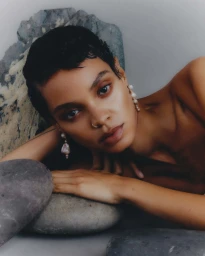 Portrait of a woman with short dark hair wearing pearl earrings resting against smooth gray rocks.