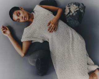 Model with short dark hair wearing a textured cream dress lying on grey studio backdrop surrounded by dark stones