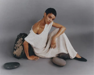 Model Akira Reid seated in a textured off-white knit dress beside smooth stones against a grey studio backdrop.