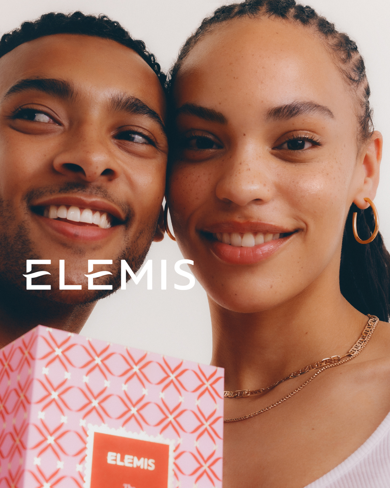Cosmetic product advertisement featuring a smiling man and woman holding an Elemis skincare gift set in a light, bright indoor setting.