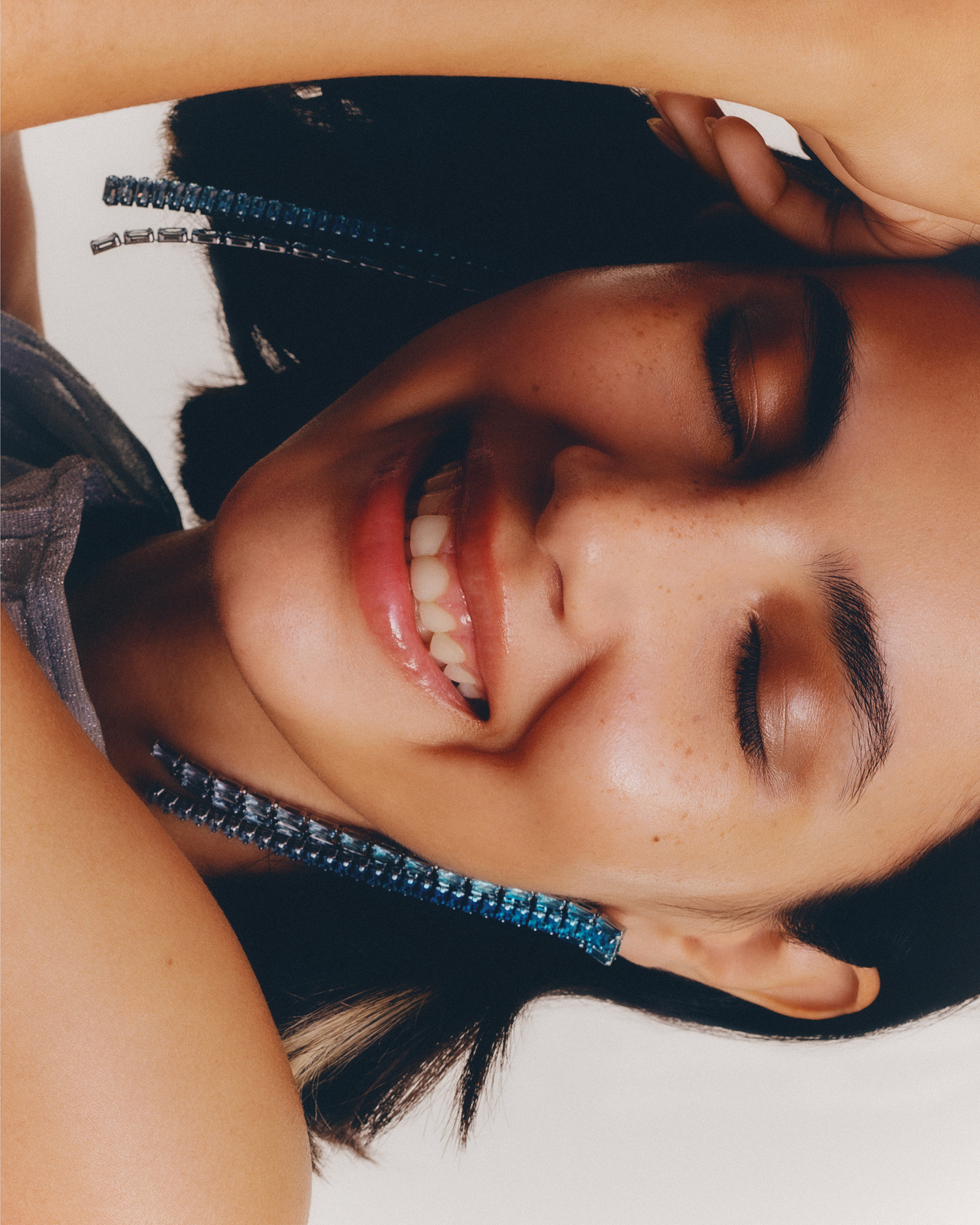 Close-up beauty shot of a woman with dark hair smiling with her eyes closed, wearing a grey top and blue linear earrings.