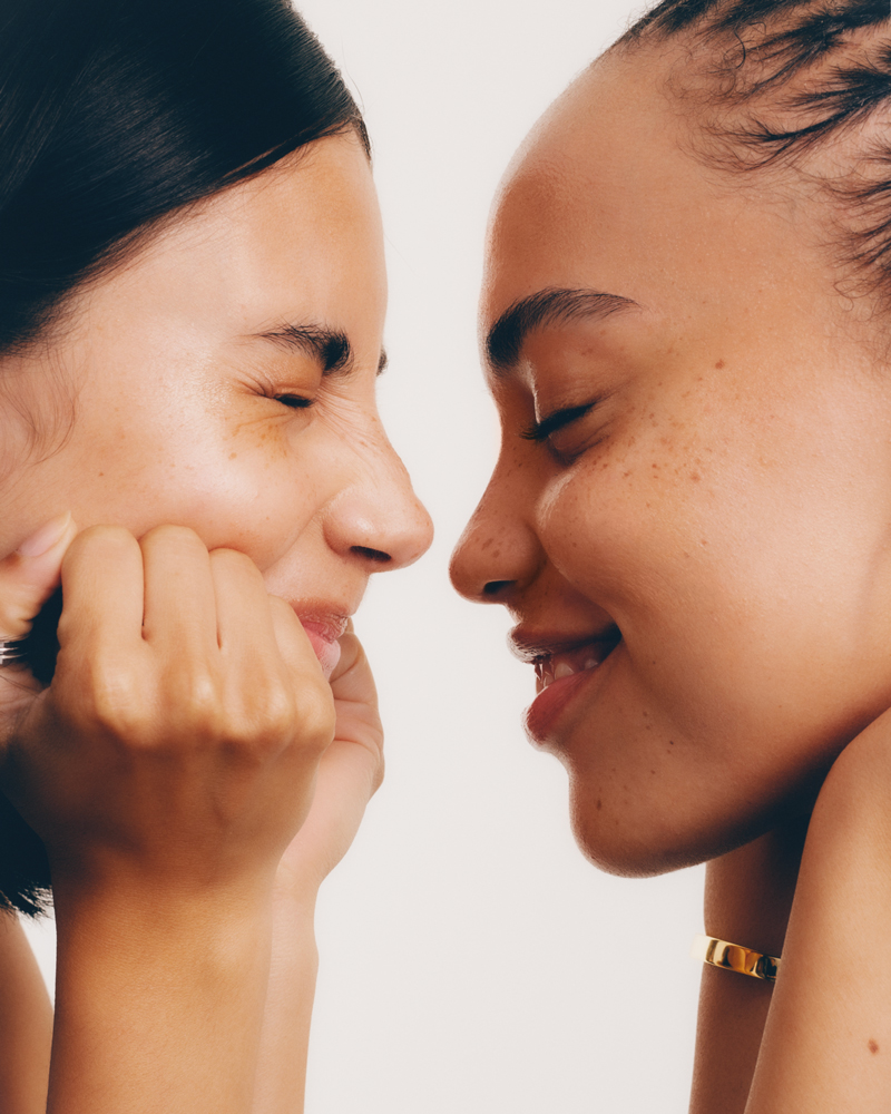 Close-up of two smiling women with fair skin and dark hair in a light studio setting. Beauty shot for skincare or cosmetic use.