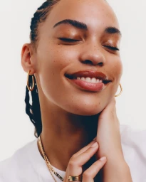 Close-up beauty portrait of a smiling woman with freckles, wearing gold hoop earrings and resting her hand on her neck against a white background.