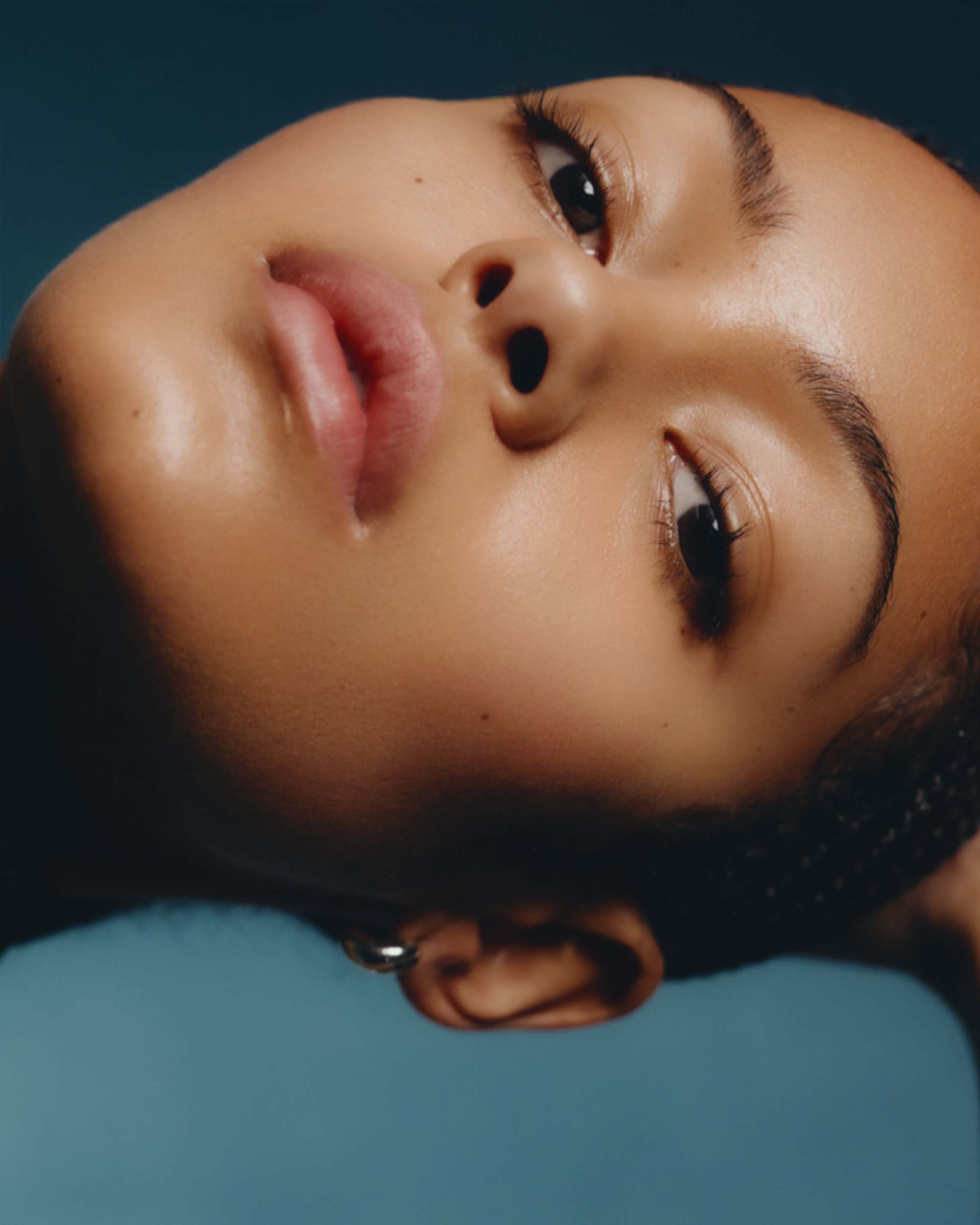 Close-up portrait of a young African American woman with glowing skin, facing upward against a teal-colored backdrop in a studio setting.