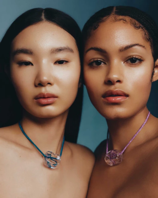 Close-up of two diverse women with clear glass pendant necklaces against a soft blue backdrop in a beauty and fashion context.