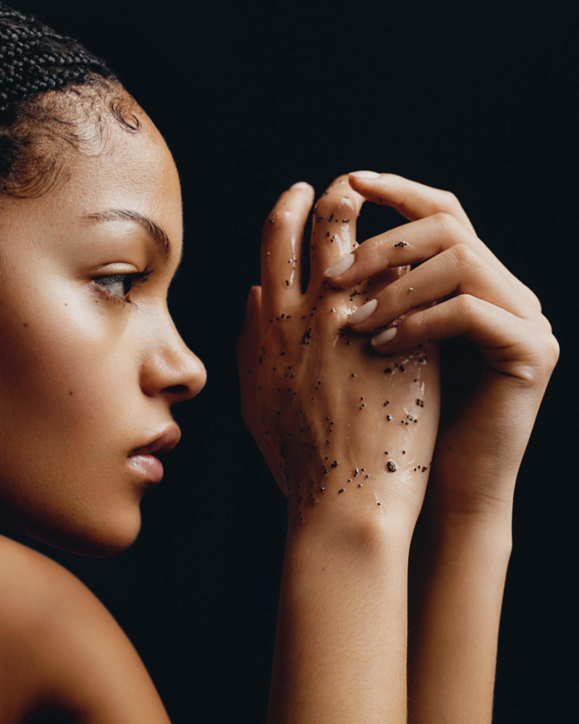 Profile of a young woman with braided hair, hands covered in dark specks against a black background in a beauty or skincare context.