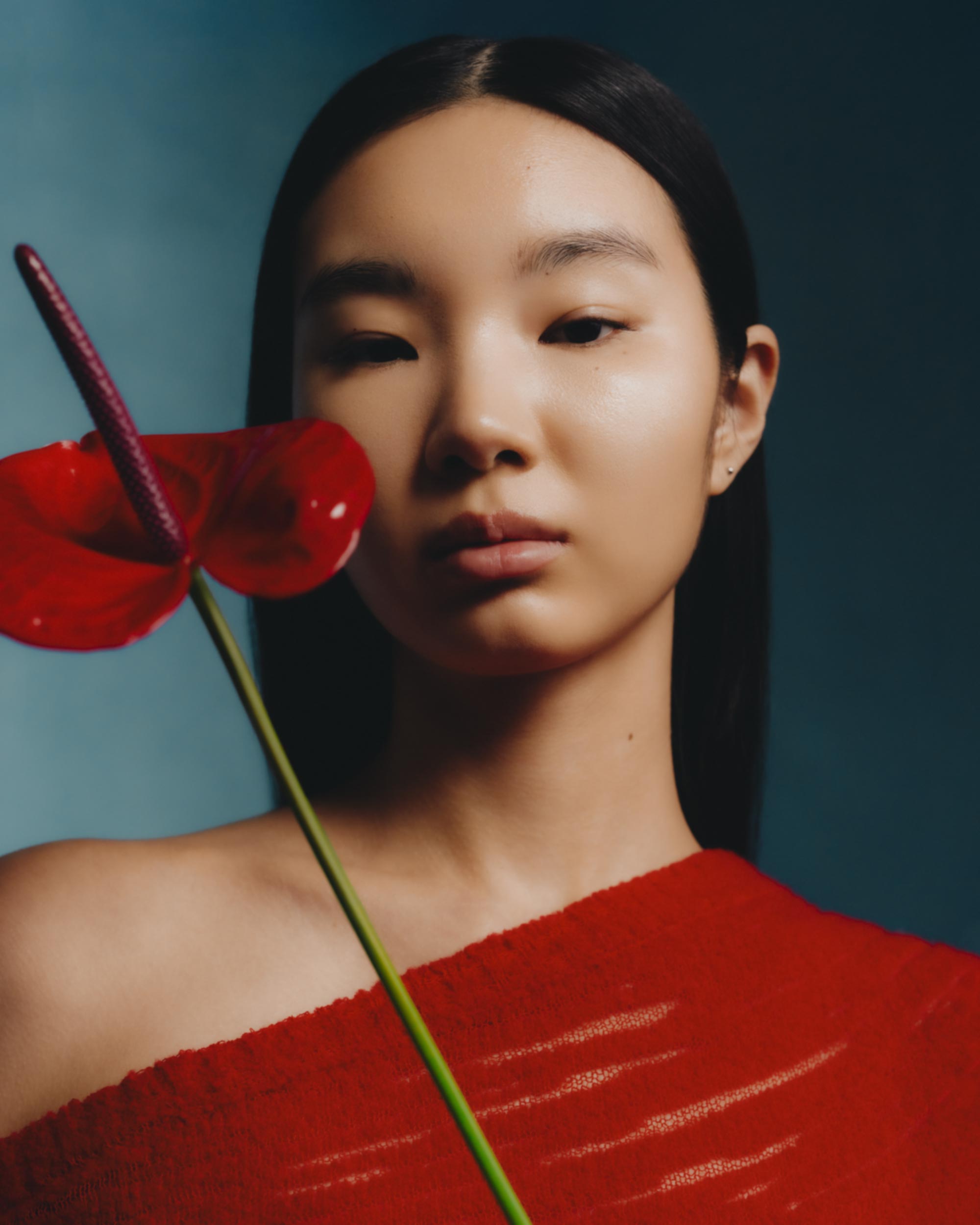 Asian woman poses with a red Anthurium flower in a studio setting. She wears a matching red top against a dark teal background.
