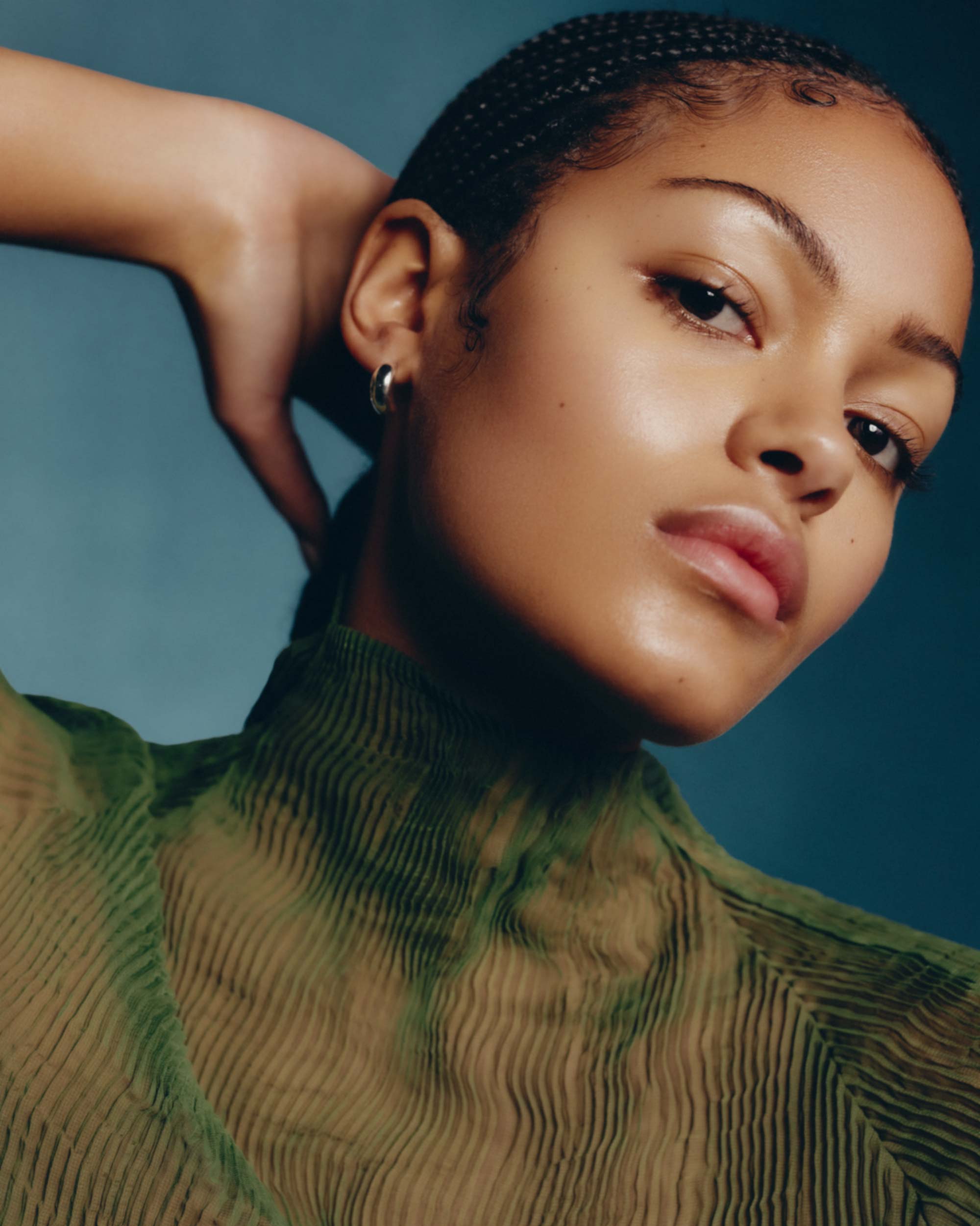 Portrait of a young adult woman with braided hair, wearing a translucent green high-neck top, posing against a dark teal backdrop.