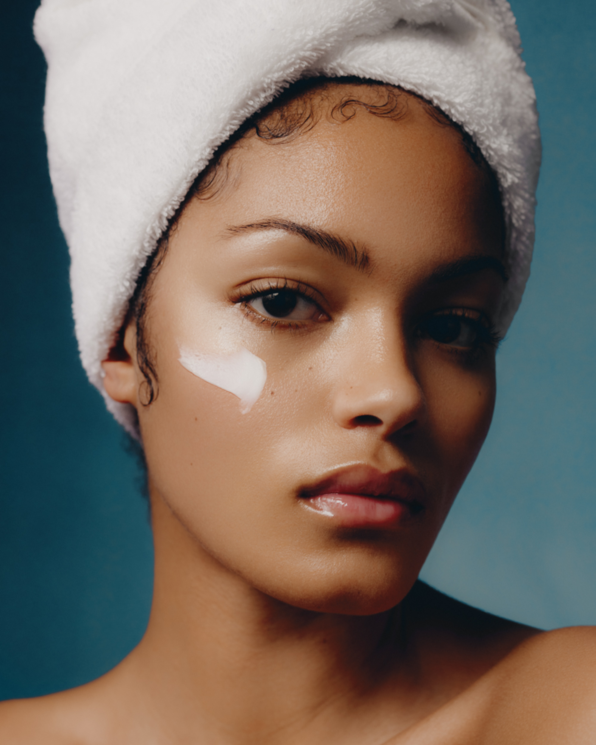 Close-up portrait of an adult female with cream on her face, wearing a white towel on her head against a blue background.