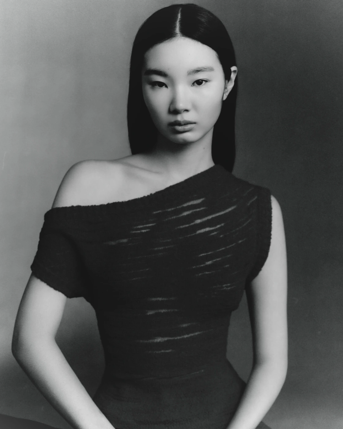 Portrait of a woman with long dark hair wearing a black one-shoulder top against a neutral gradient studio background.