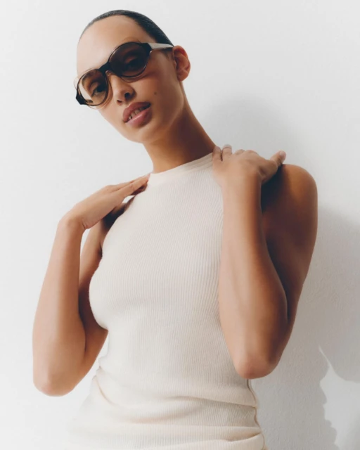 Woman in ribbed top wears sunglasses against white backdrop, portrait format, eye-level angle, showcases fashion and beauty.