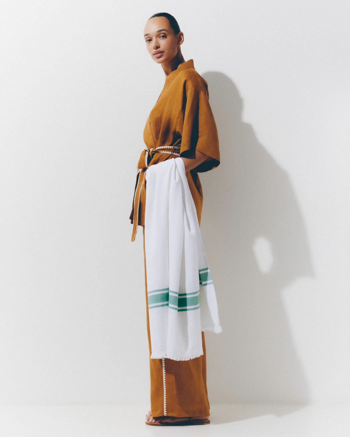 Woman in a brown kimono-style robe poses in a brightly lit photo studio with a white and green striped towel over her arm.