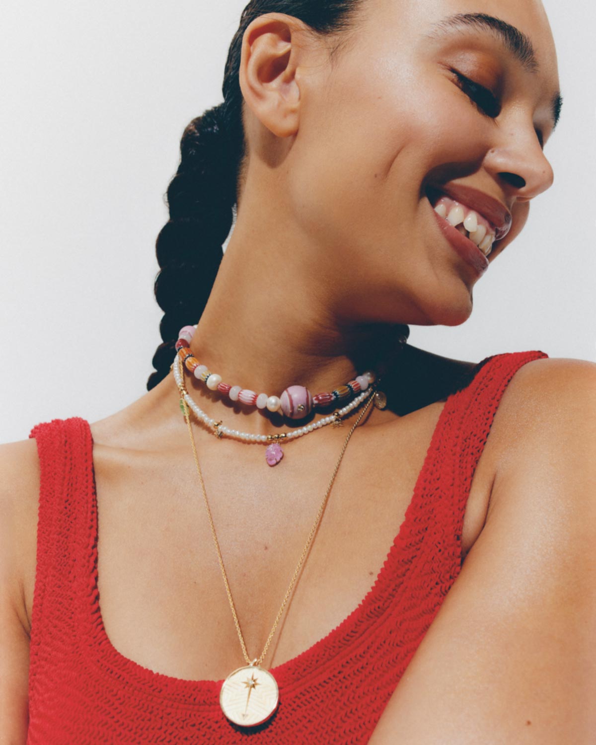 Close-up of an adult woman with braided hair smiling and wearing colorful beaded and gold necklaces against a white background.