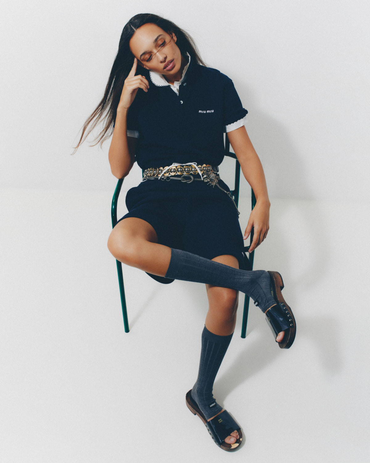 Woman in black polo shirt and shorts sitting on green chair in a studio setting. She is wearing high socks and sandal shoes.