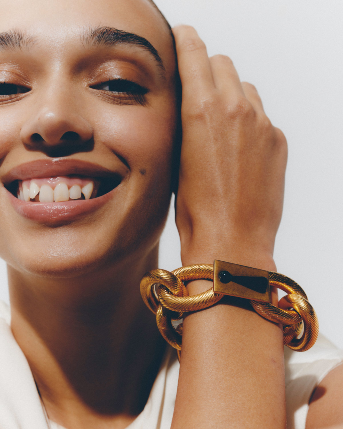 Close-up of a smiling adult female with hand raised, wearing a gold bracelet on her wrist against a plain white background.