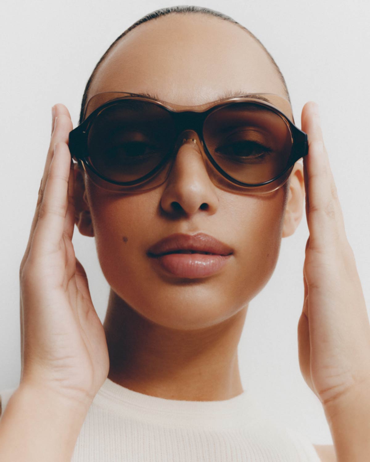 Close-up portrait of a woman wearing oversized tinted sunglasses with hands raised to her temples against a plain white studio background.