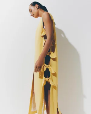 Model in profile wearing a long sleeveless yellow dress with bow-tied side cutouts against a white studio wall.