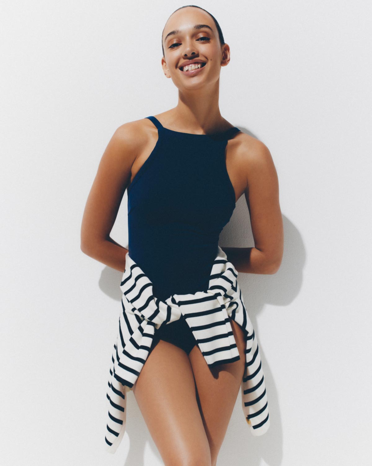 Model smiling in a dark navy one-piece swimsuit with a striped sweater tied around her waist against a bright white background.