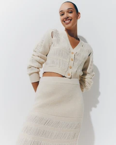 Smiling model wearing a textured cream knit cardigan and matching skirt against a plain white seamless studio background.