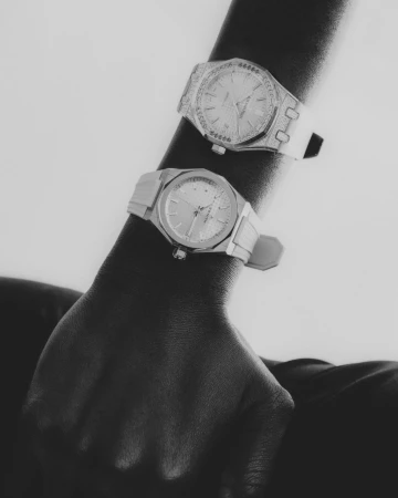 Close-up studio shot of two wristwatches on an arm, highlighting their dials, metal bracelets, and overall sleek design in black and white.