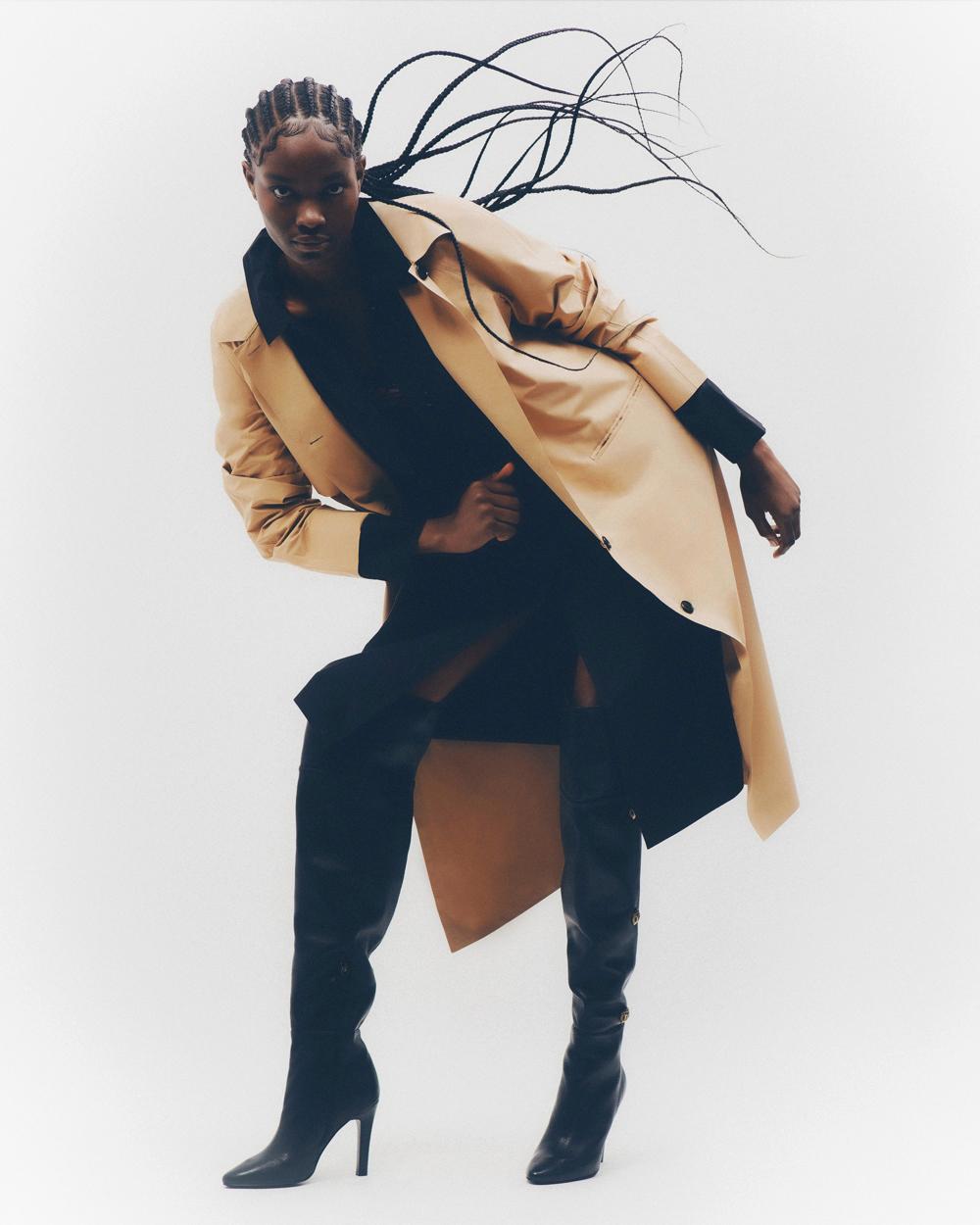 A young Black woman poses in a studio wearing a trench coat, black top and thigh-high heeled boots against a white background.