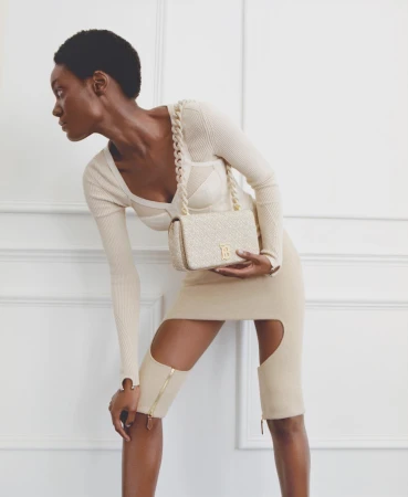 A slender Black woman poses in a beige dress with a handbag against a simple white wall background, capturing a minimalist and fashionable aesthetic.