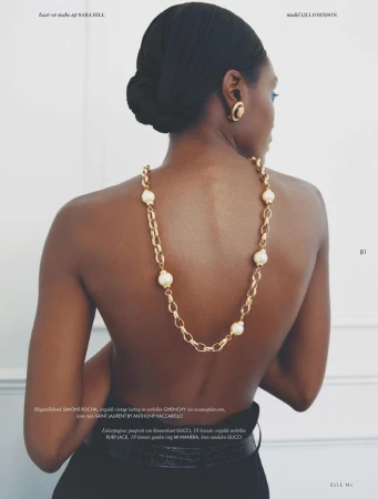 Black woman from behind wearing a gold chain necklace with large pearl accents draped down her bare back against a white wall.