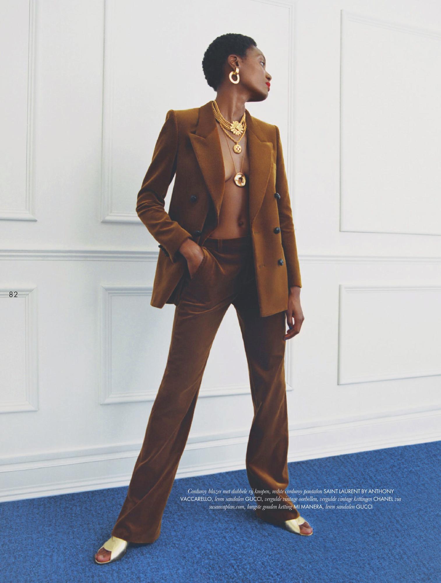 Fashion editorial shot: Black female model poses in a brown velvet blazer and pants with gold jewelry indoors against a white wall and blue carpet.