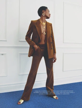 Fashion editorial shot: Black female model poses in a brown velvet blazer and pants with gold jewelry indoors against a white wall and blue carpet.