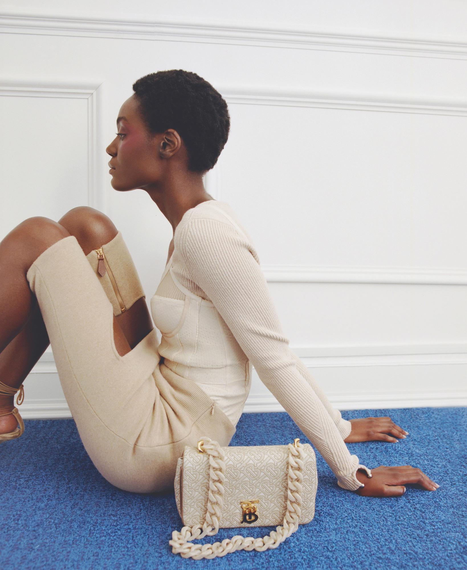 Elegant dark-skinned woman poses with a Burberry purse in an indoor studio, wearing a matching knit outfit and blue carpet beneath her.