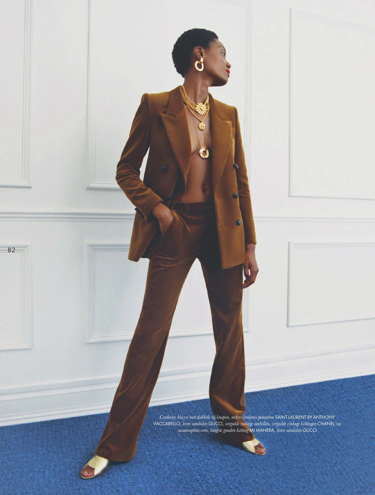 Model in brown velvet pantsuit with layered gold necklaces standing on a blue carpet against white wall panels
