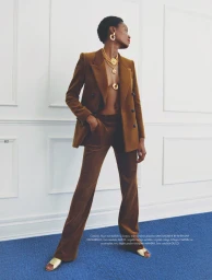 Model in brown velvet pantsuit with layered gold necklaces standing on a blue carpet against white wall panels