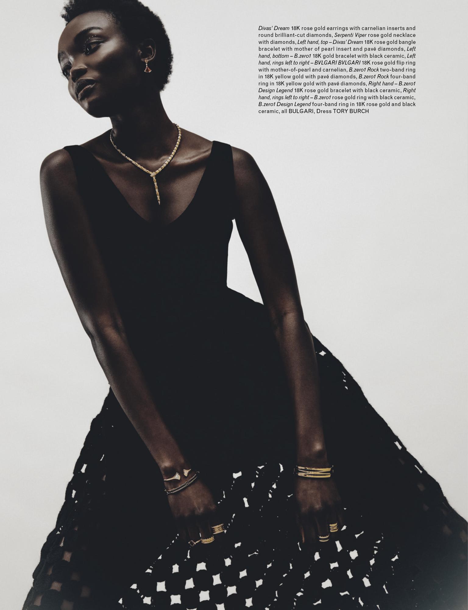 Elegant black woman poses in black V-neck dress with open crochet-style skirt, adorned with gold earrings, necklace, bangles, and rings.