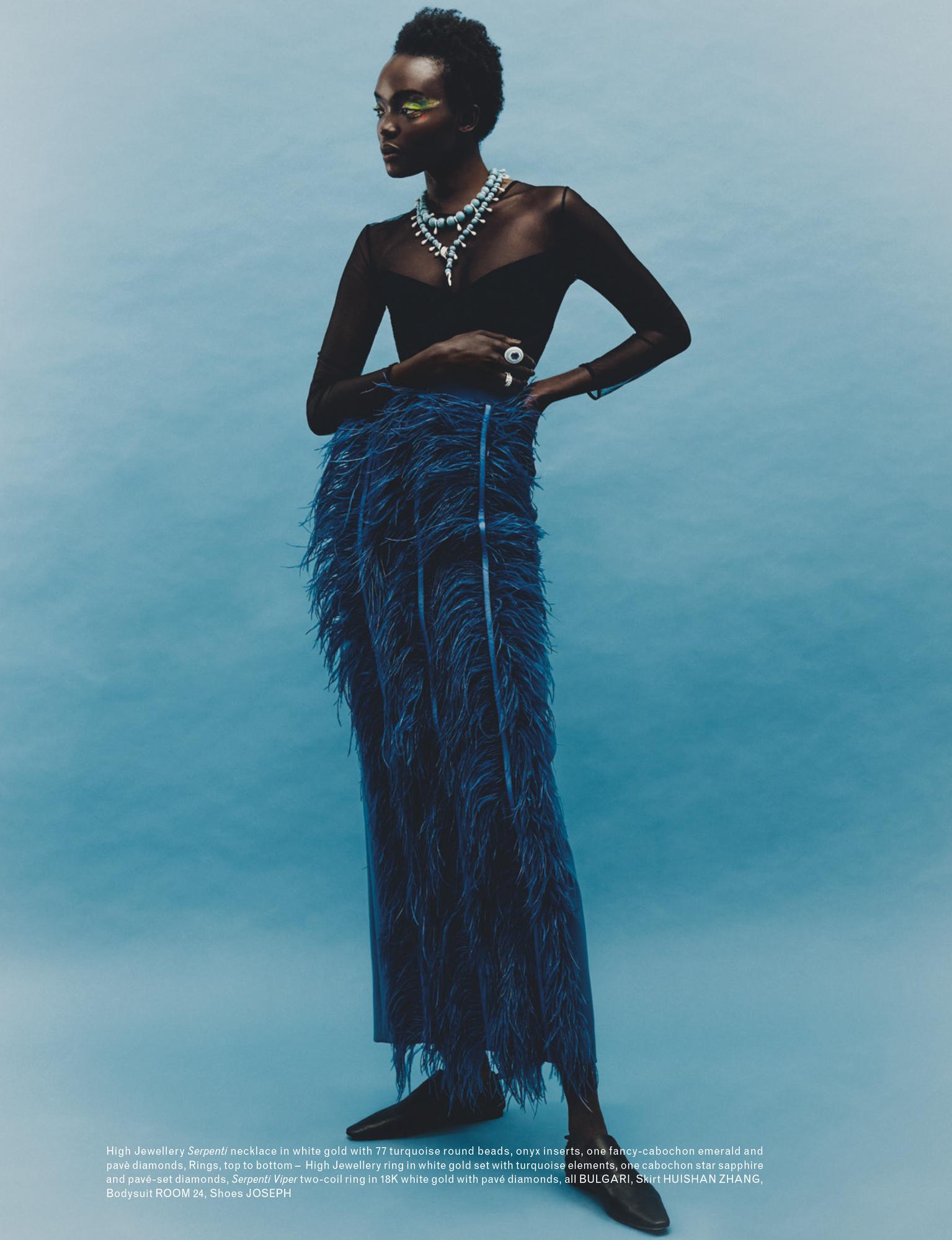 Elegant woman posing in a long-sleeved black top and long navy feather skirt, accessorized with jewelry, against a blue background.