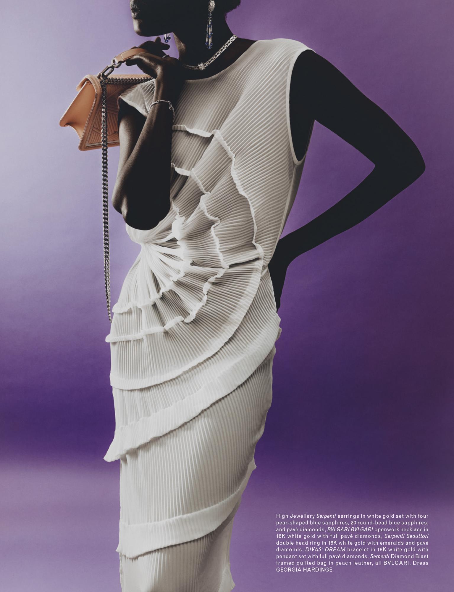 A fashion portrait features a Black woman wearing a white pleated dress, silver jewelry, and a peach handbag against a purple backdrop.