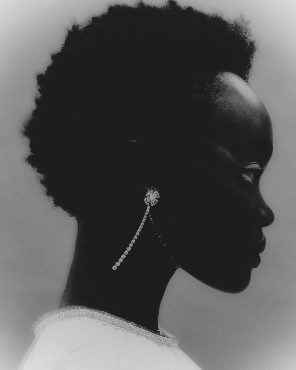 Profile portrait of an adult woman with dark skin and natural hair, wearing a white top and sparkling earring, against a soft gray background.