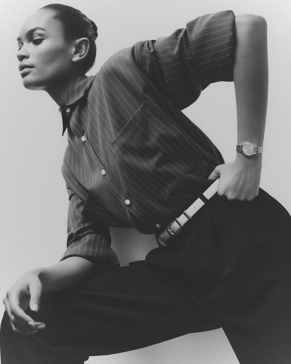 A woman poses in a pinstripe shirt and wide-leg trousers, accented by a white belt and watch, set against a neutral backdrop in monochrome.