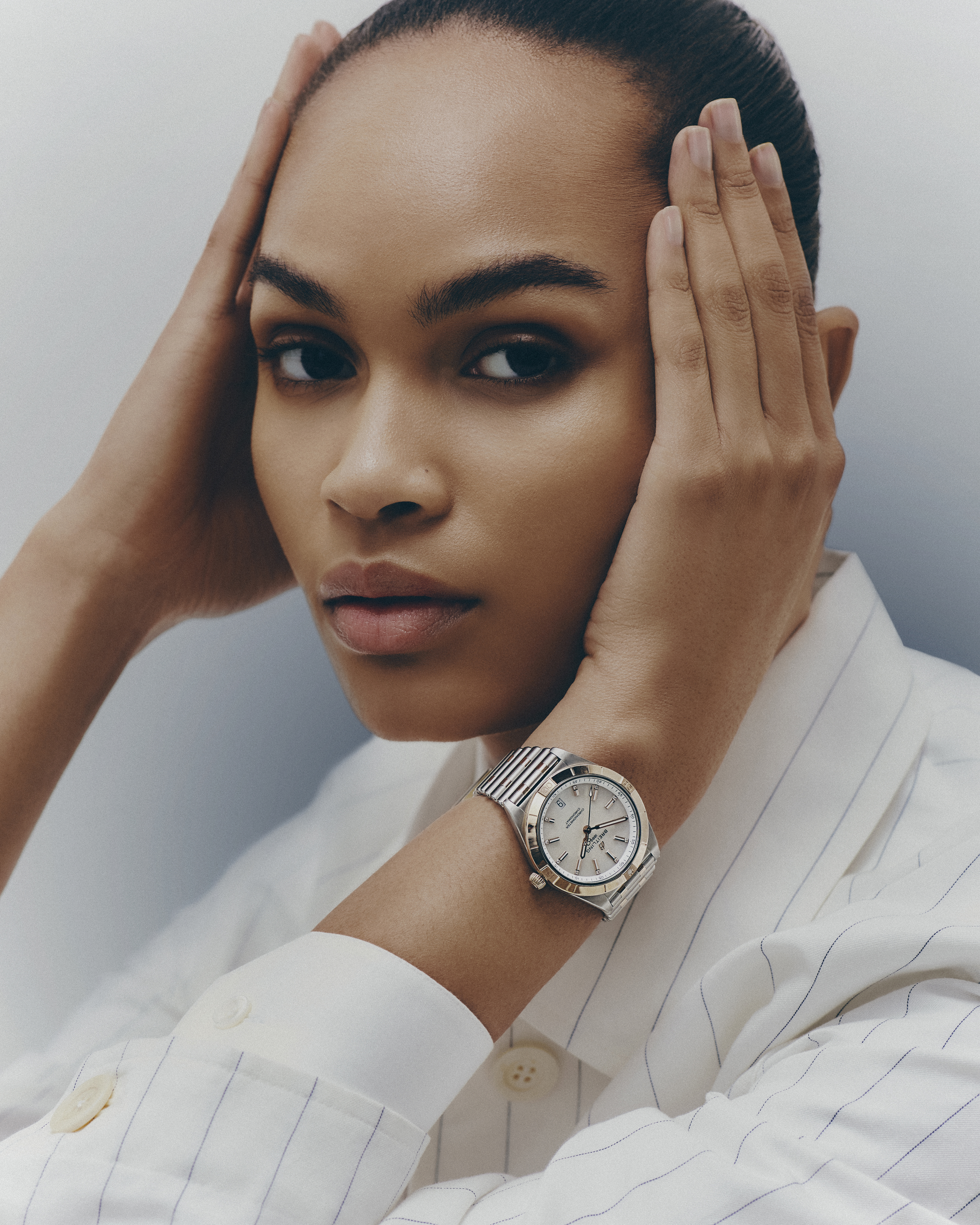 Elegant portrait of an adult woman with her hands near her face and wearing a white striped blazer and silver wristwatch in a photo studio.