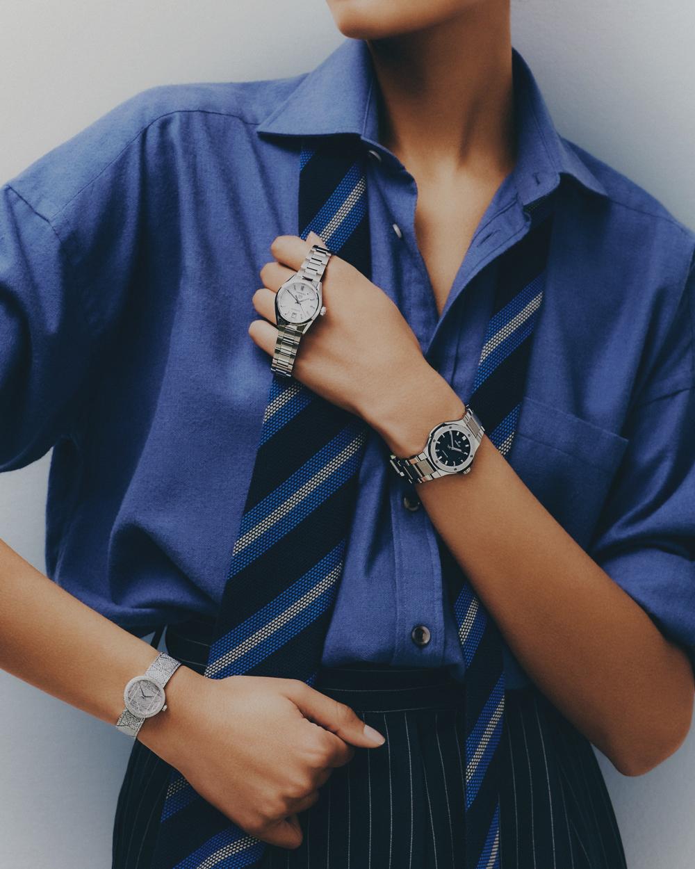 Close-up of a woman's torso with two wristwatches on her left arm and one on her right, set against a blue collared shirt and striped tie.