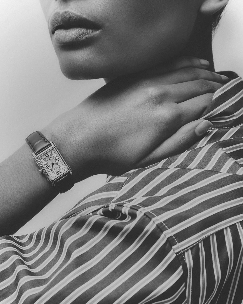 Black and white close-up of an adult female's arm and wrist with a classic rectangular wristwatch, wearing a striped shirt. Minimalist shot.